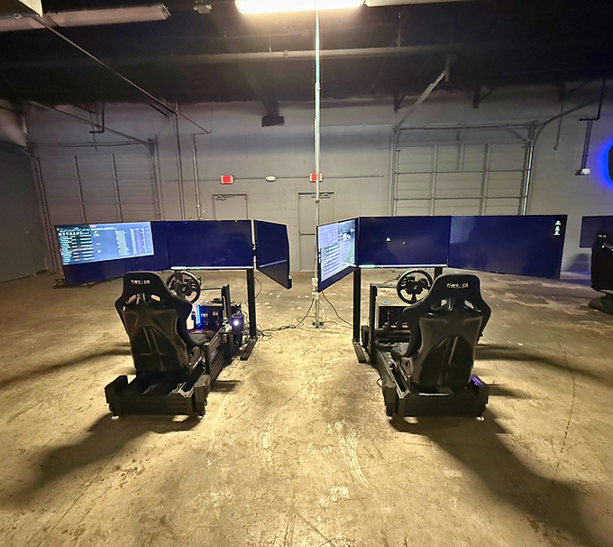 Two racing simulators in a warehouse, each with a large curved screen and racing seat setup.