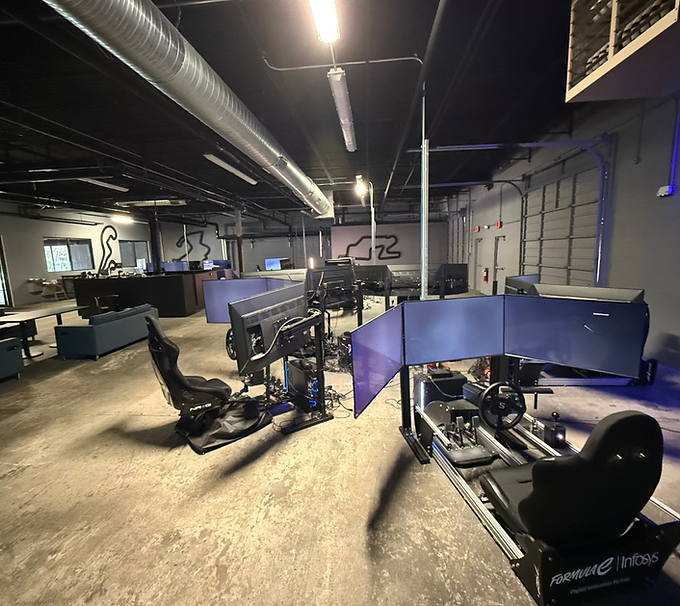 Modern office with racing simulators, dual monitors, and blue privacy panels under industrial ceiling lights