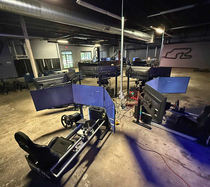 Indoor racing simulator room with multiple black cockpit rigs and monitors under industrial lighting