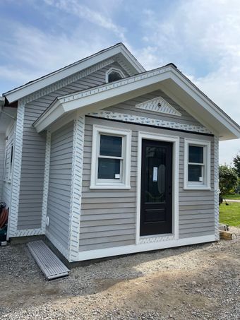 Small gray house with white trim and black front door on a gravel lot
