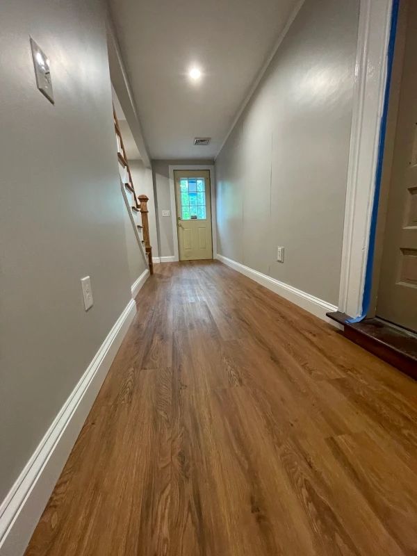 Narrow hallway with wood floors, gray walls, and a bright doorway at the far end.