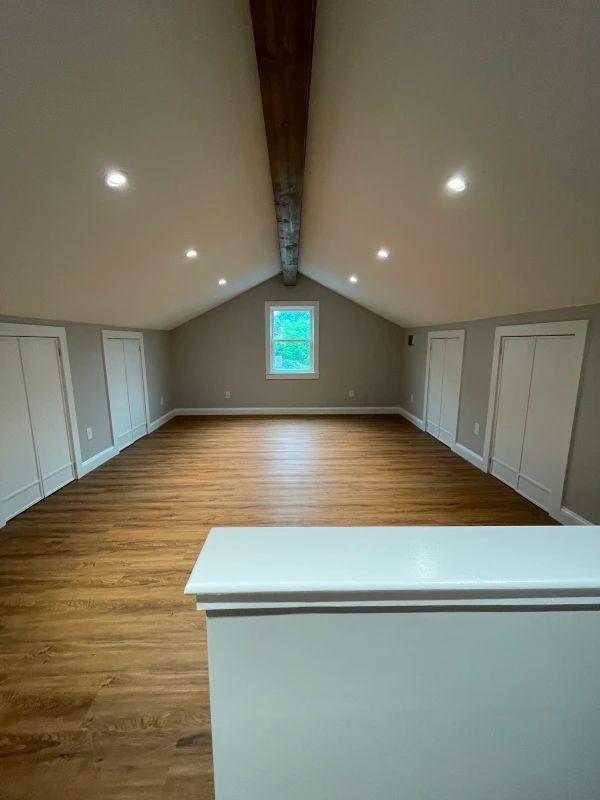 Empty finished attic room with wood floors, white walls, recessed lights, and a central window.