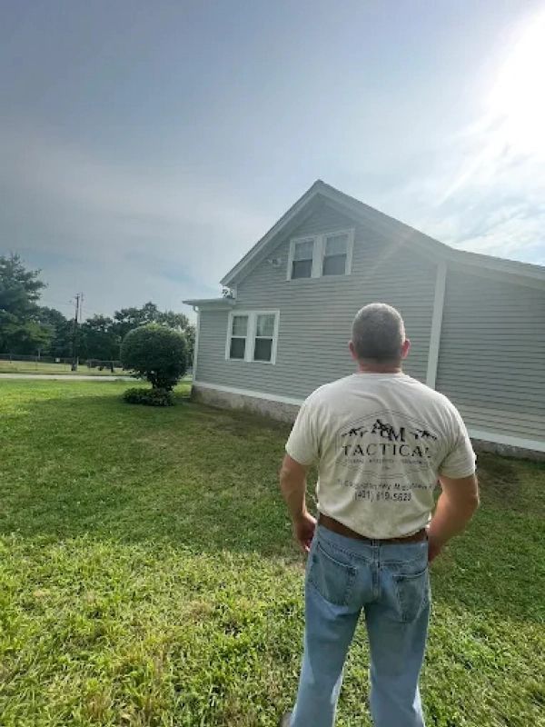 Person in a white T-shirt and jeans stands on a grassy lawn facing a gray house in bright sunlight.