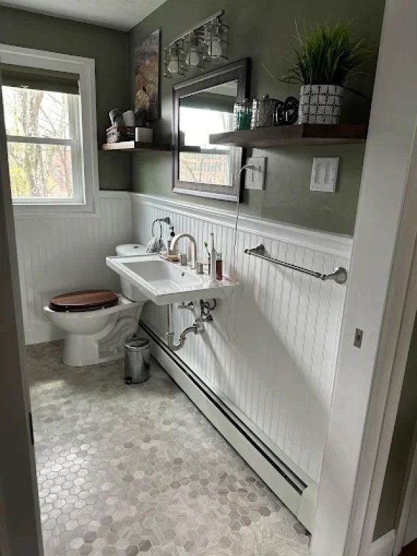 Small gray bathroom with pedestal sink, toilet, open shelves, and white beadboard wainscoting