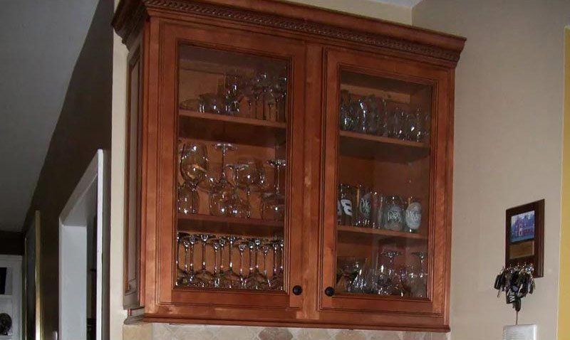 Wooden wall cabinet with glass doors displaying glassware and stemware in a home interior
