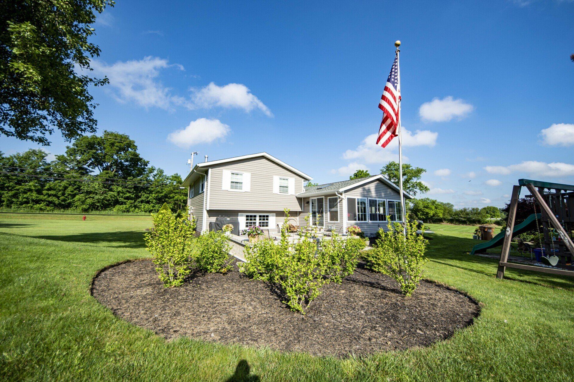 House with American flag, surrounded by green grass and landscaping on a sunny day.