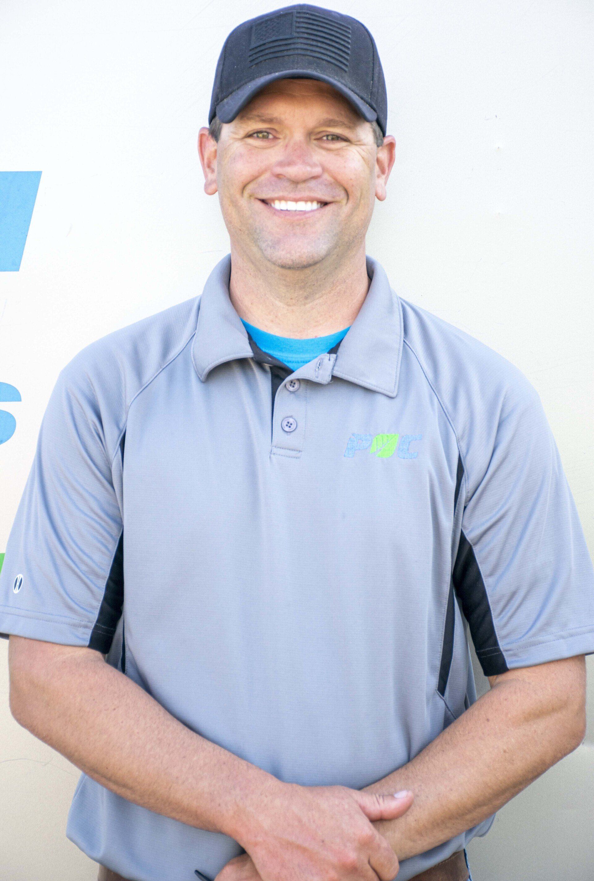 Man in gray polo shirt and baseball cap smiling, arms crossed.