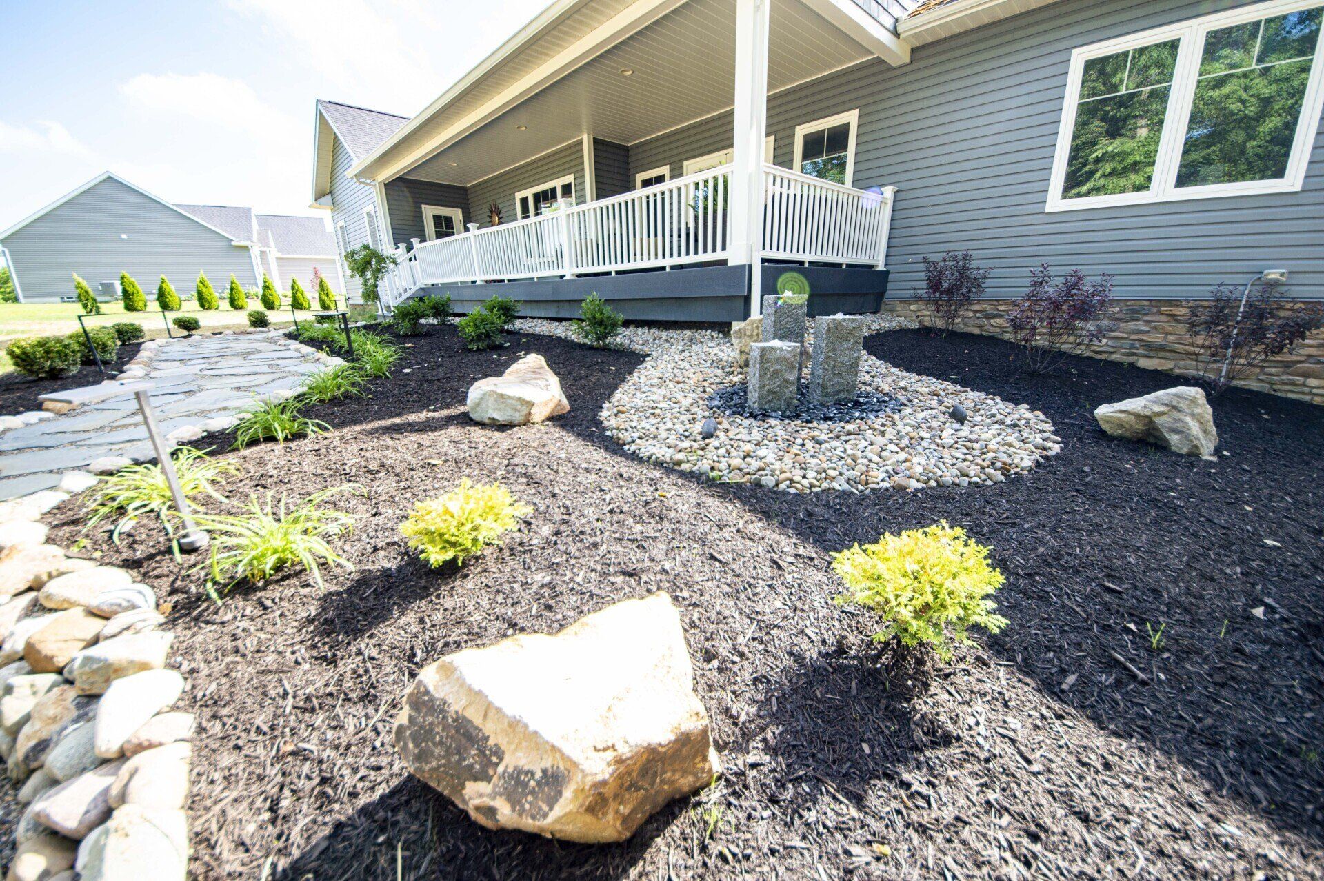 Landscaped yard with black mulch, rocks, and small plants in front of a gray house with a porch.