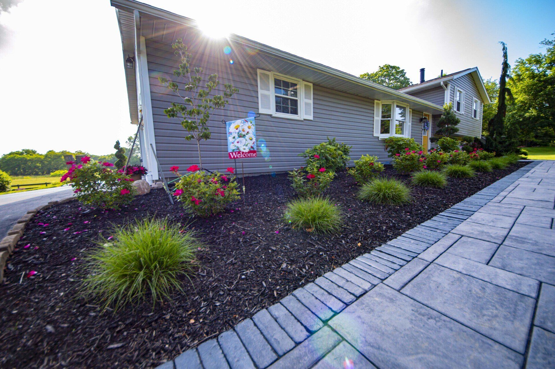 Gray house with landscaped flowerbeds and a stone walkway in sunlight.