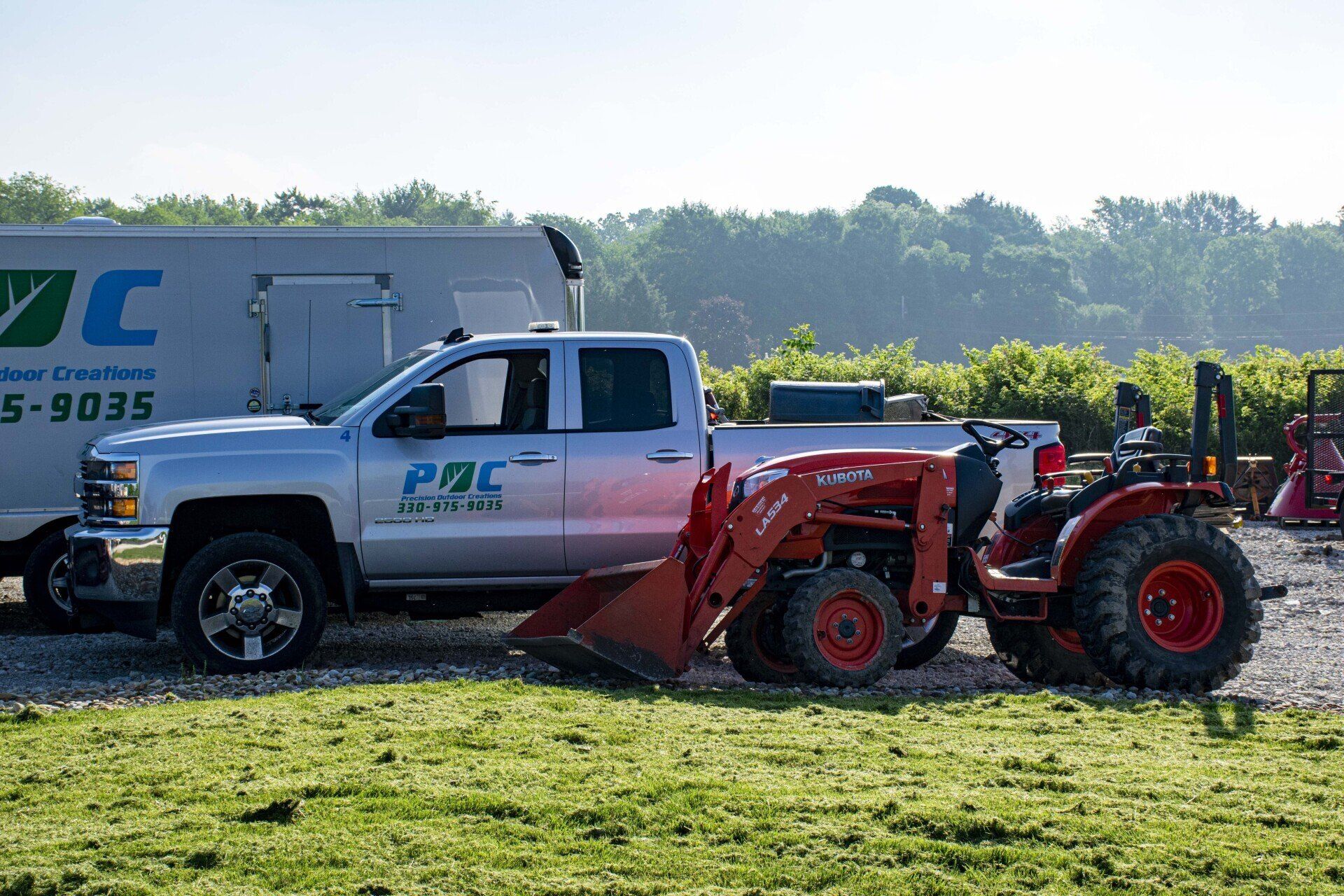 Silver truck with a red tractor on a trailer, parked on grass near a building and vineyard.