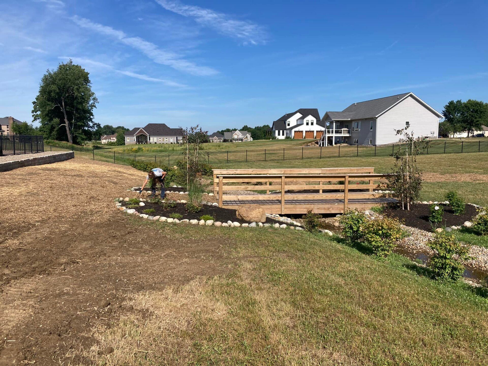 Wooden bridge over a creek in a grassy area, with houses in the background under a blue sky.