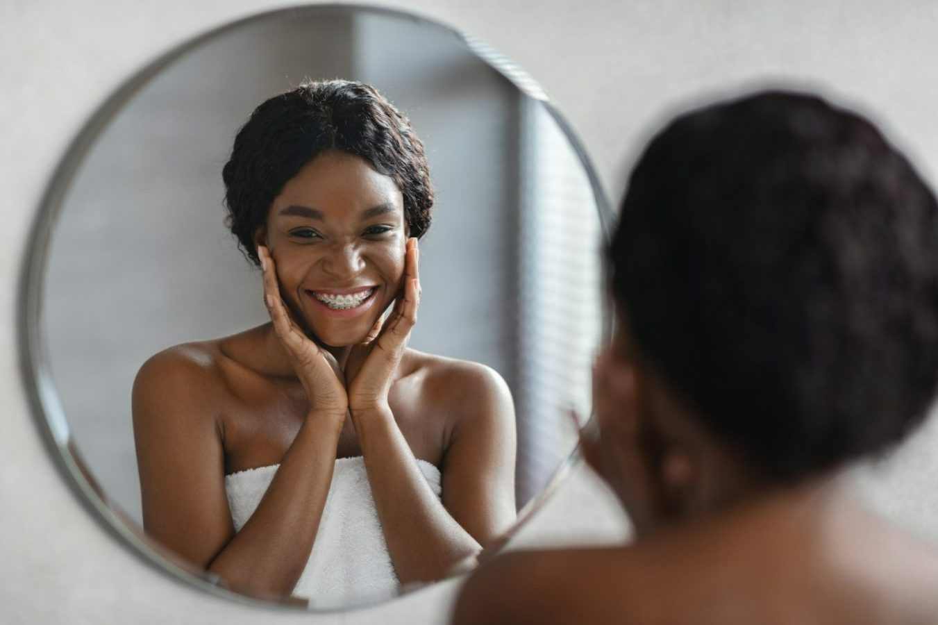 Woman smiles, touching her face in a mirror, wrapped in a white towel.