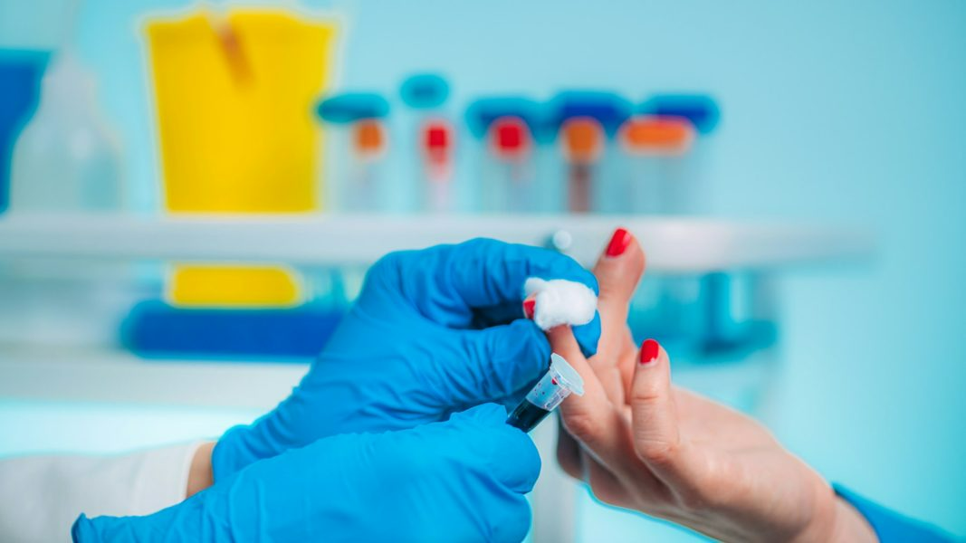 Person in blue gloves drawing blood from a finger in a medical setting.