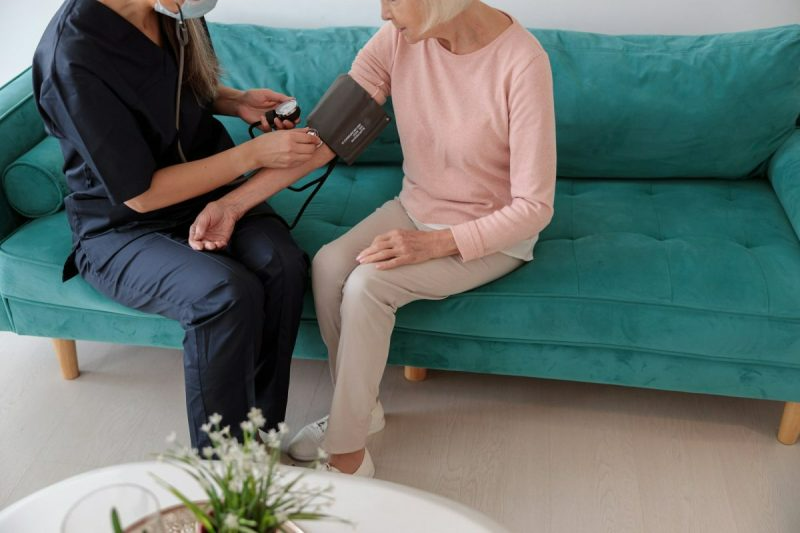 A healthcare worker taking a patient's blood pressure on a turquoise sofa.
