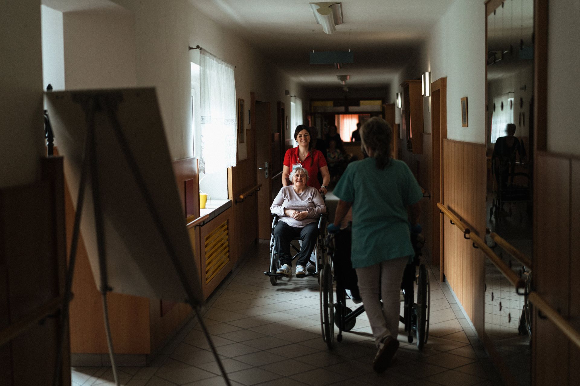 People walking with walkers in a dim hallway, with a wheelchair in the center.