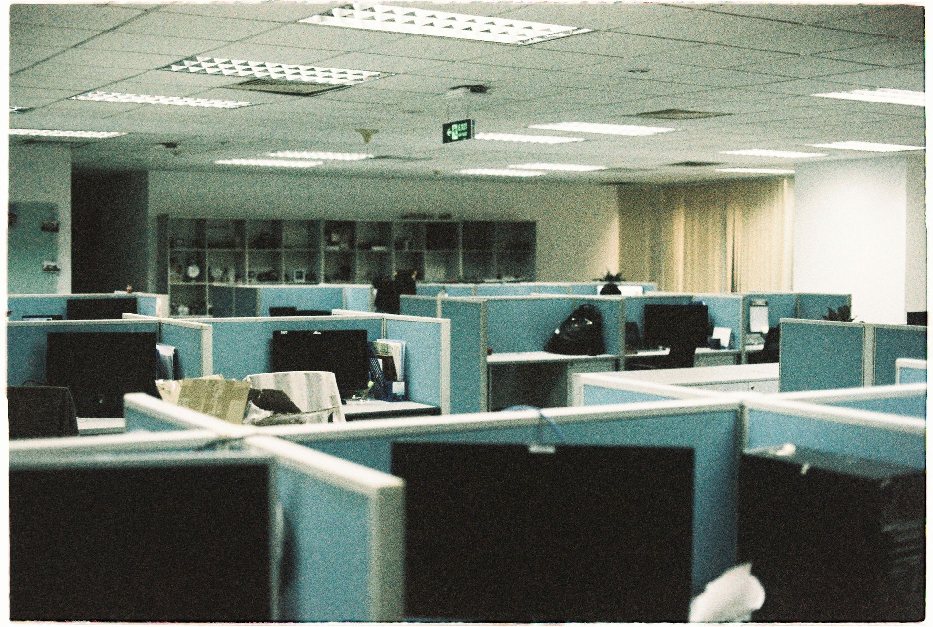 Empty office cubicles in a dimly lit workspace with teal partitions and fluorescent ceiling lights
