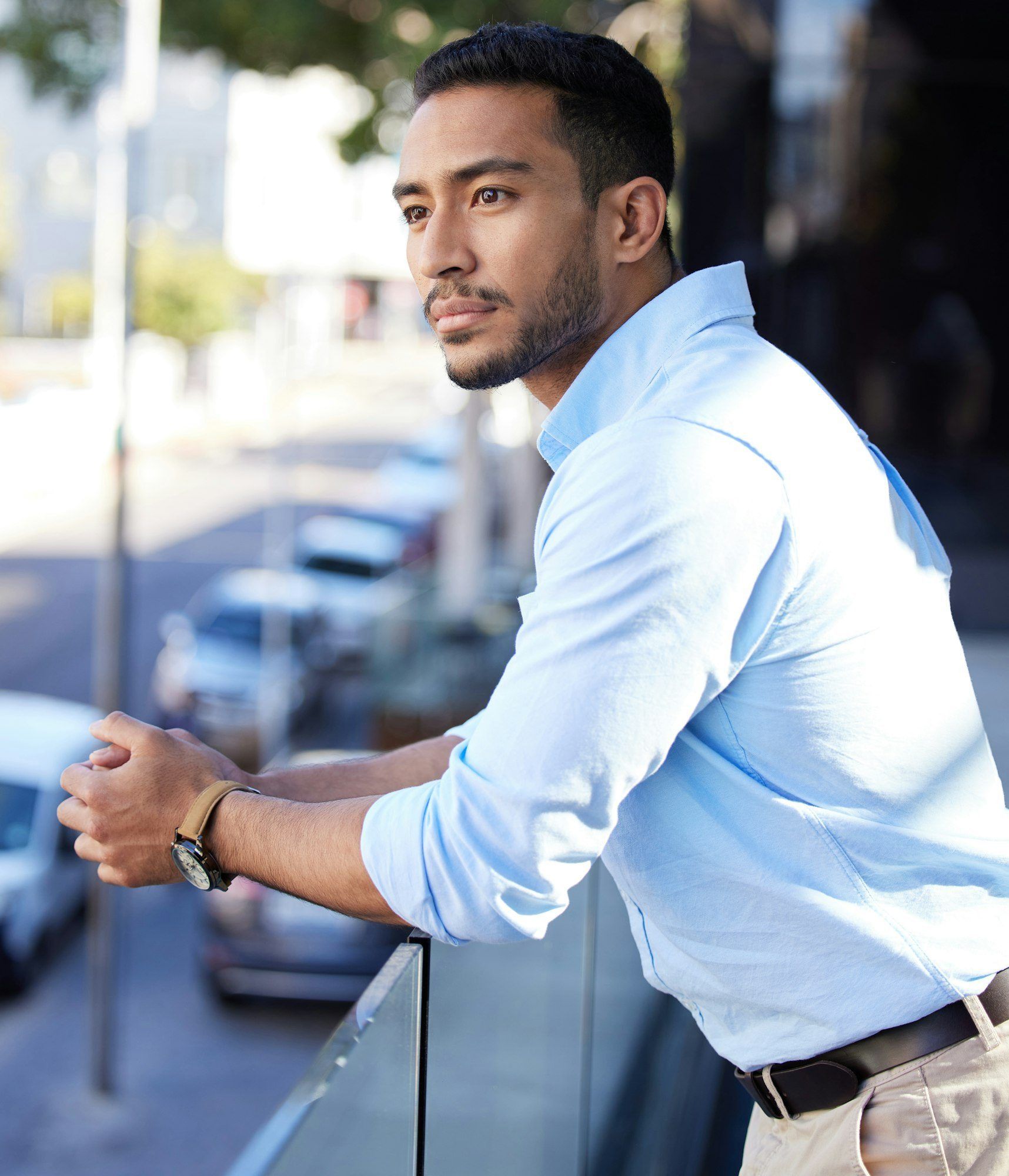 Man in blue shirt, leaning on a balcony, looking thoughtful, city backdrop.