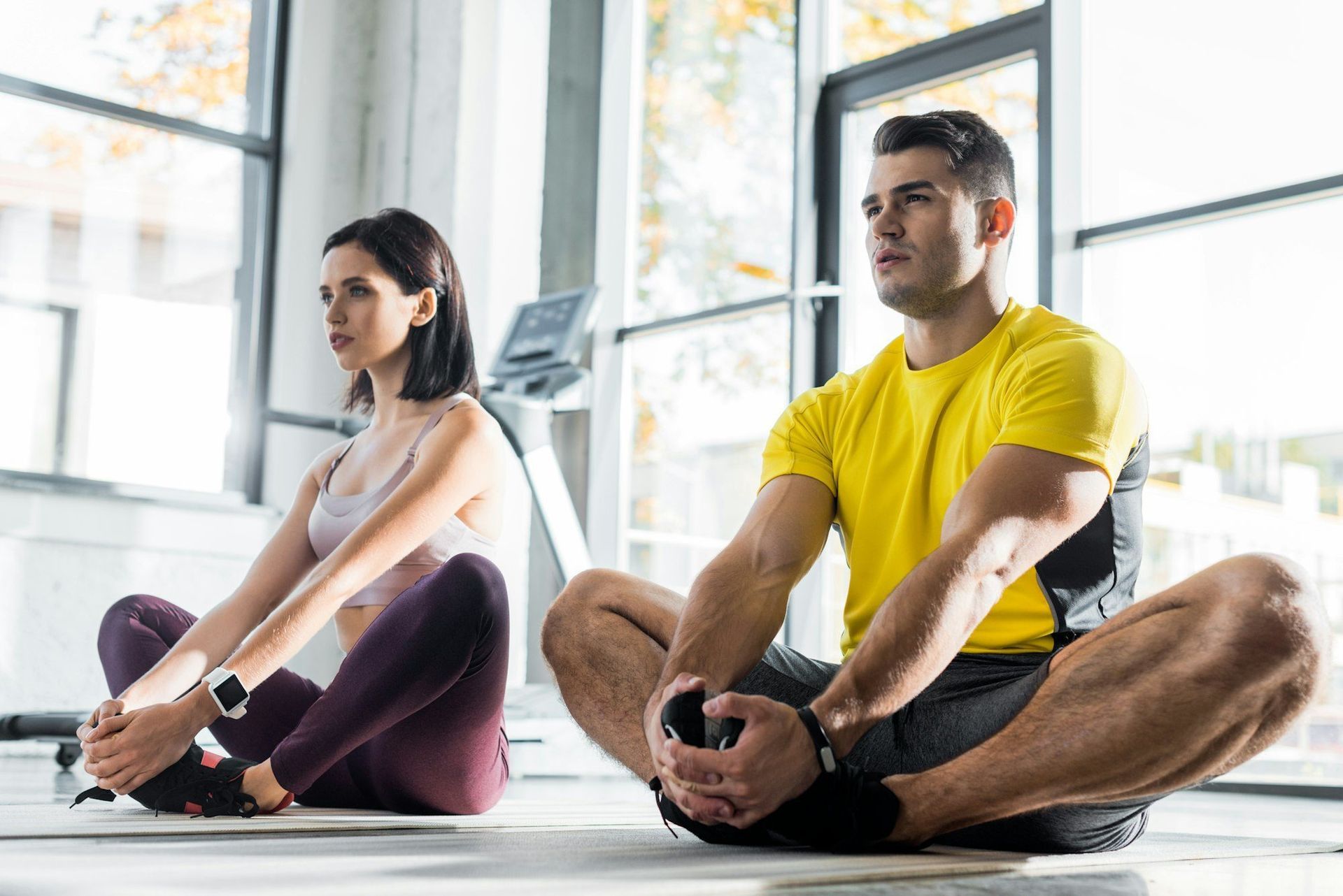 Two people stretching in butterfly pose on yoga mats in a gym.