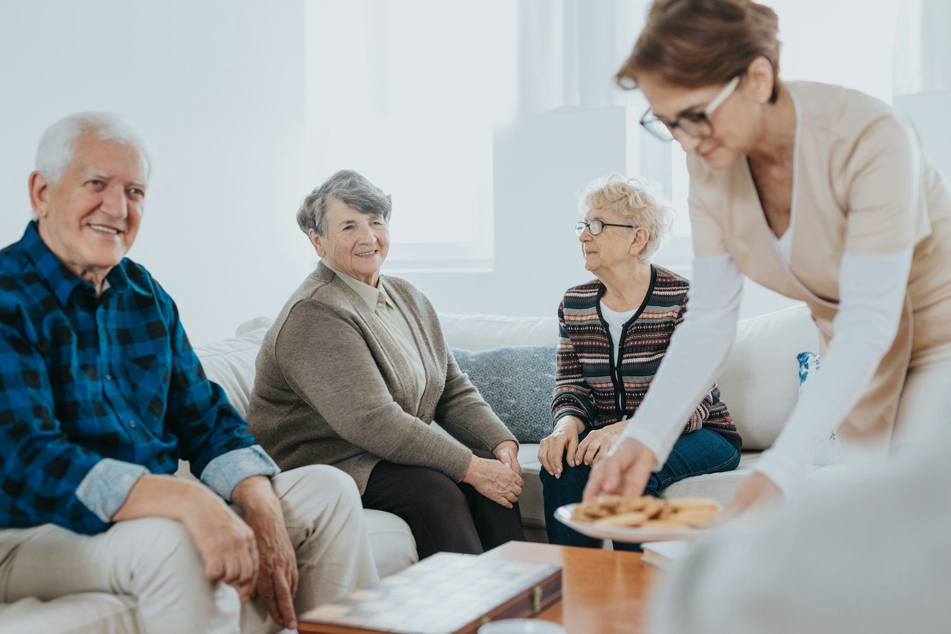 Four seniors on a couch, a caregiver offers a plate of cookies.