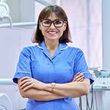 A female dentist is standing in a dental office with her arms crossed and smiling.