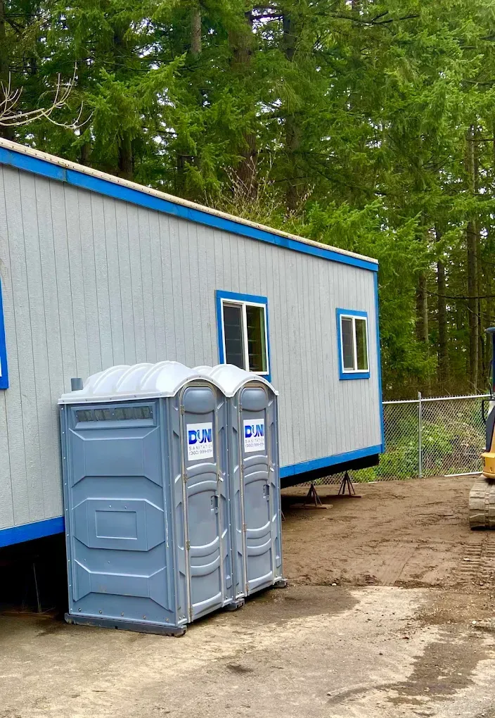 Portable toilets beside a blue-gray construction trailer in a wooded outdoor site