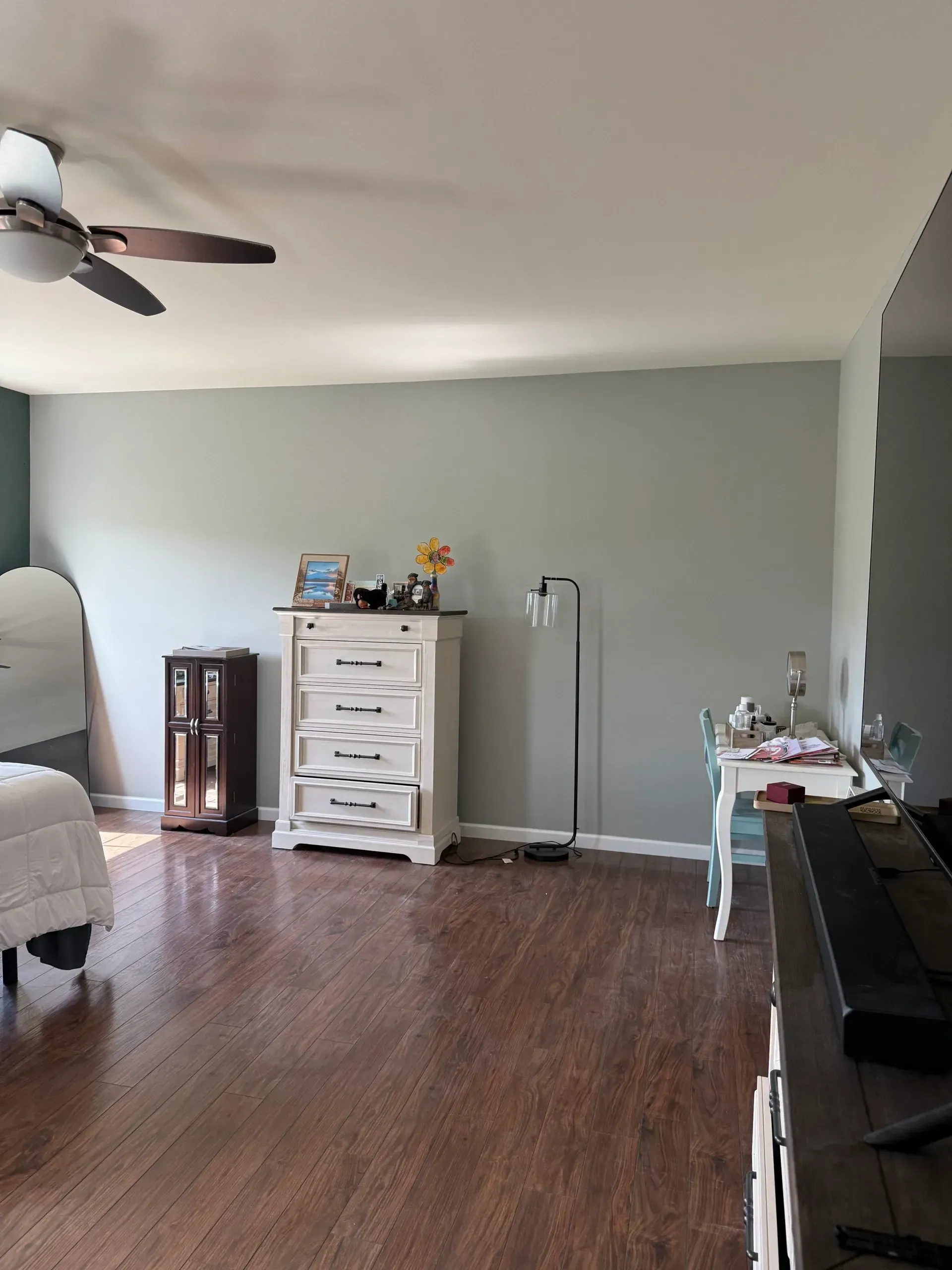Empty living room with hardwood floors, white dresser, ceiling fan, and TV stand against pale walls.