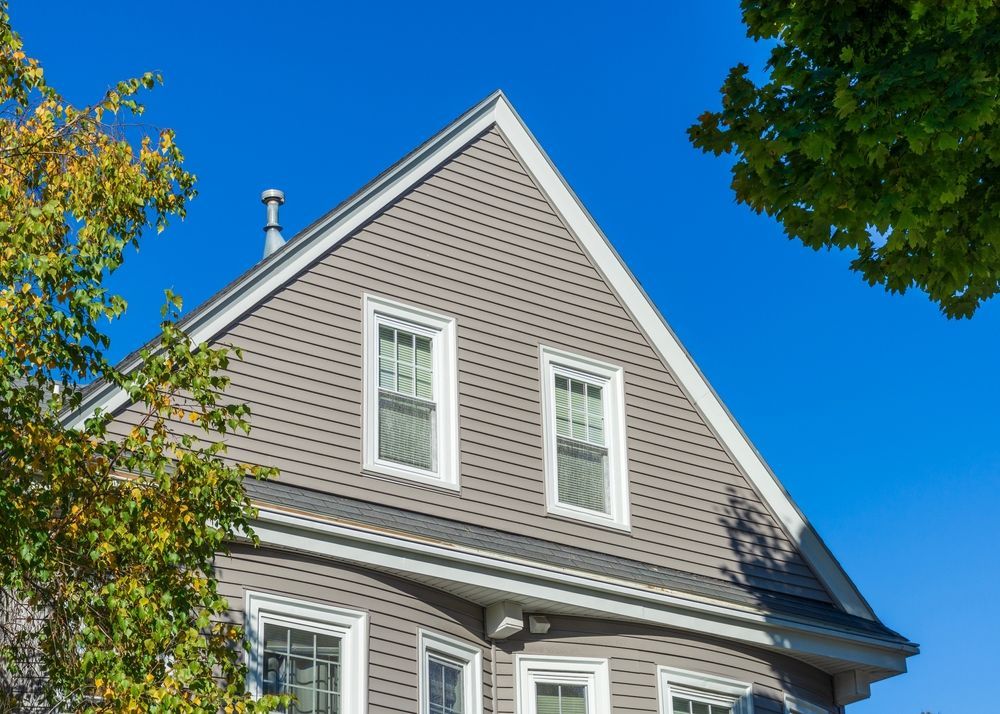 Gray house gable with white-trimmed windows against a bright blue sky, framed by tree branches