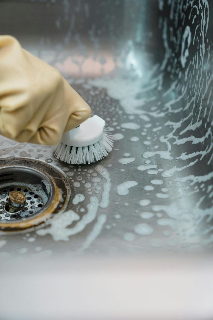Gloved hand scrubbing a soapy stainless steel sink with a round brush.