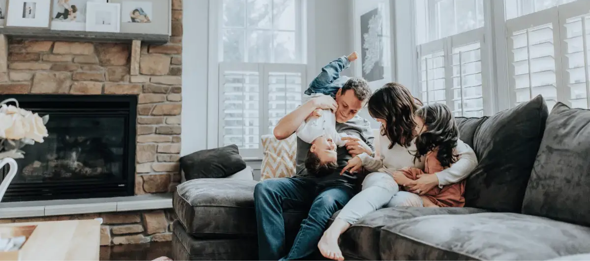 Family cuddling on a gray couch by a fireplace, smiling and playing with a baby in a bright living room