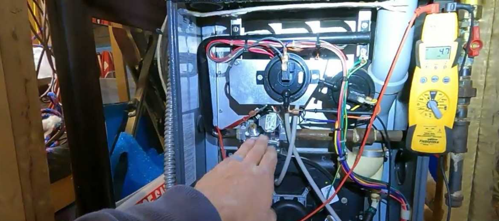 Hand pointing at a pump assembly inside a wiring-filled equipment compartment, with yellow meter on the right