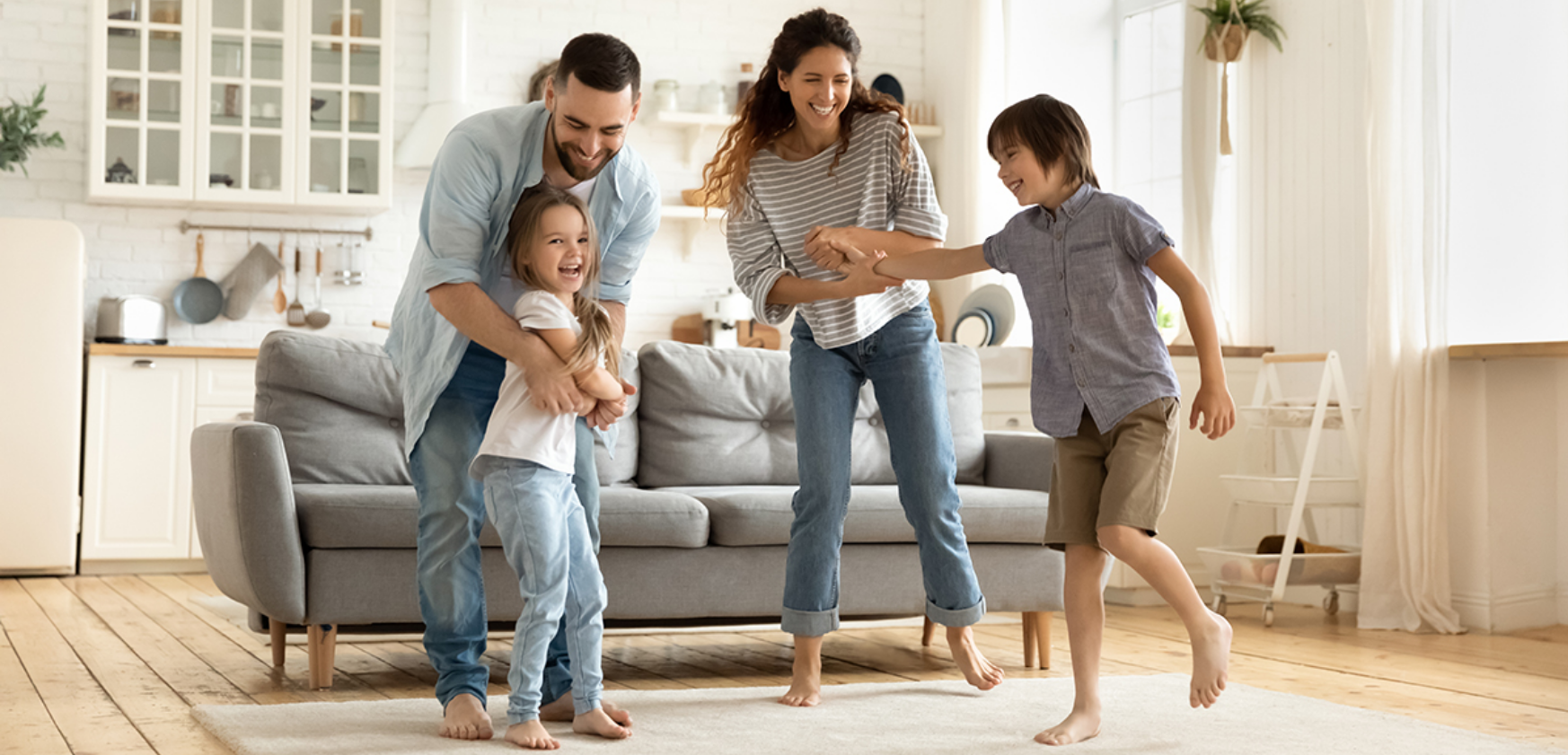 Family dancing together in a bright living room, smiling beside a gray sofa.