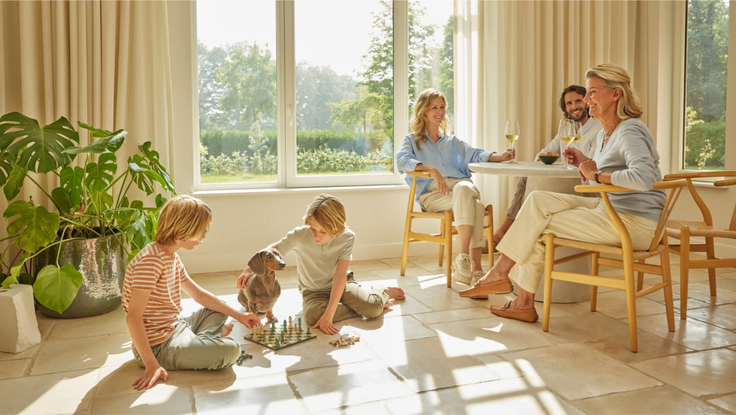 Family gathered in a sunlit living room, chatting and playing on the floor near large windows.