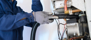 Technician in blue uniform using a vacuum hose to service machinery with exposed wires and pipes