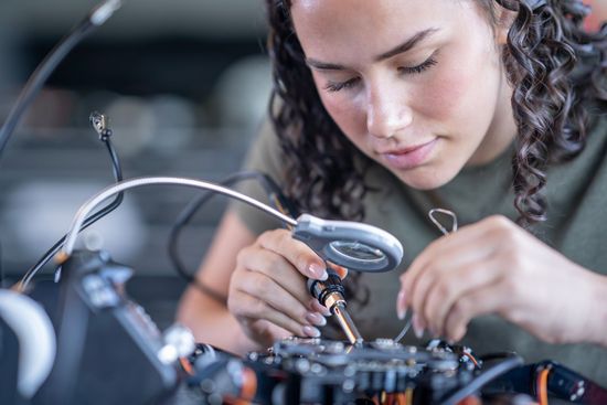 Technician closely inspecting electronics with a magnifying lamp and tools in a workshop