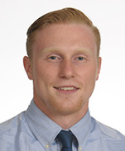 Portrait of a smiling person in a light blue shirt and dark tie against a white background