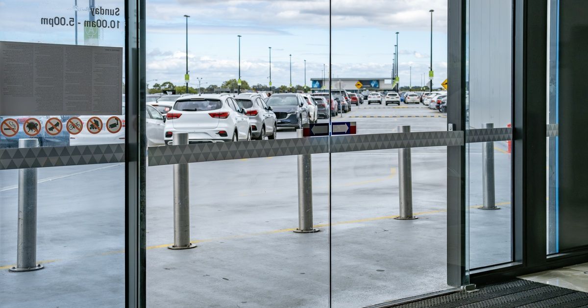 A glass shopping center entrance showing safety decals, metal bollards, and cars parked outside unde
