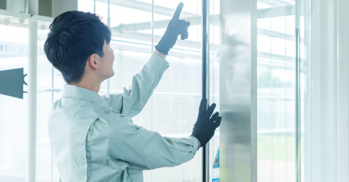 A worker in gloves pointing toward a glass automatic door while standing in a bright entrance.