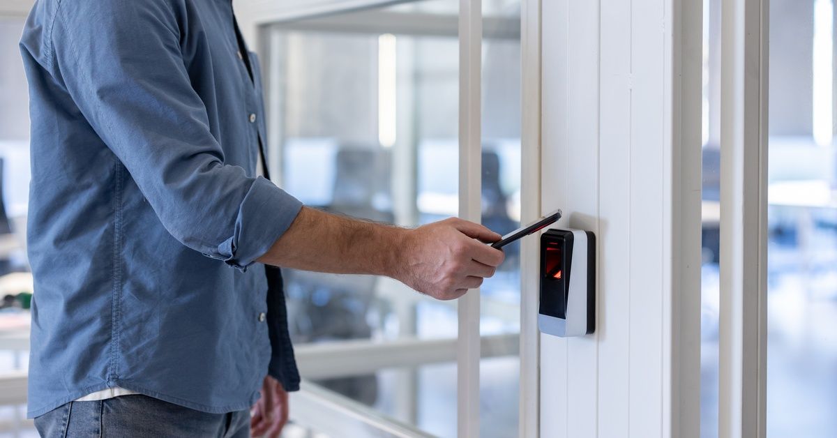 A man in a blue shirt holding a smartphone near a wall-mounted access scanner beside a glass office 