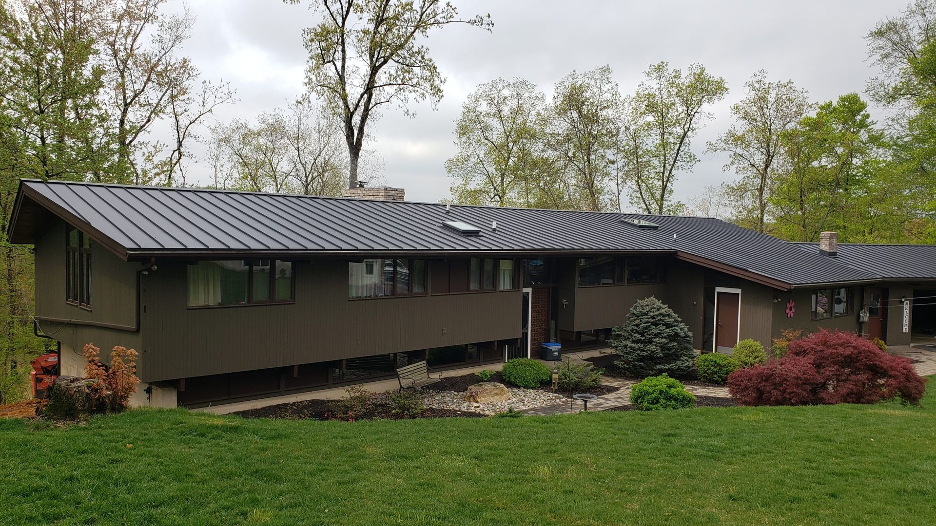 Brown mid-century modern house with dark metal roof; trees in background, green lawn in front.