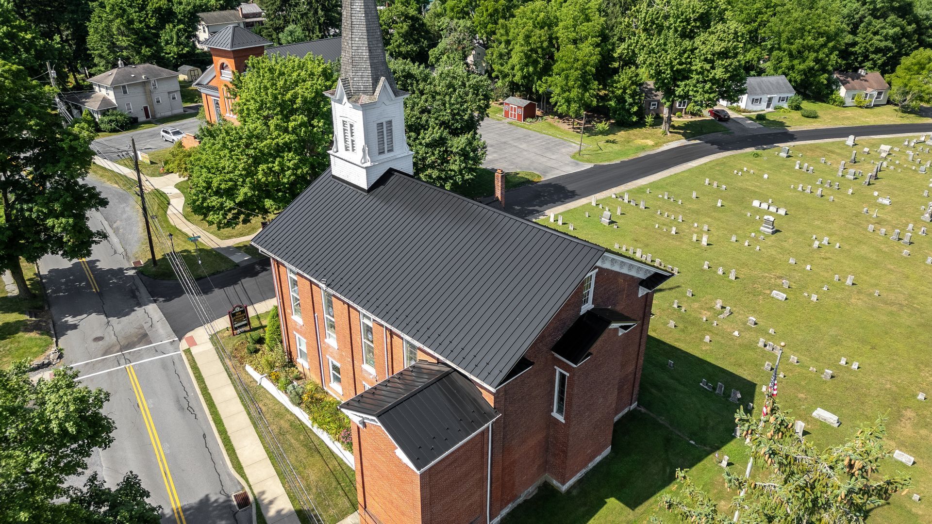 Brick church with white steeple and black roof, beside a cemetery and road, surrounded by trees.