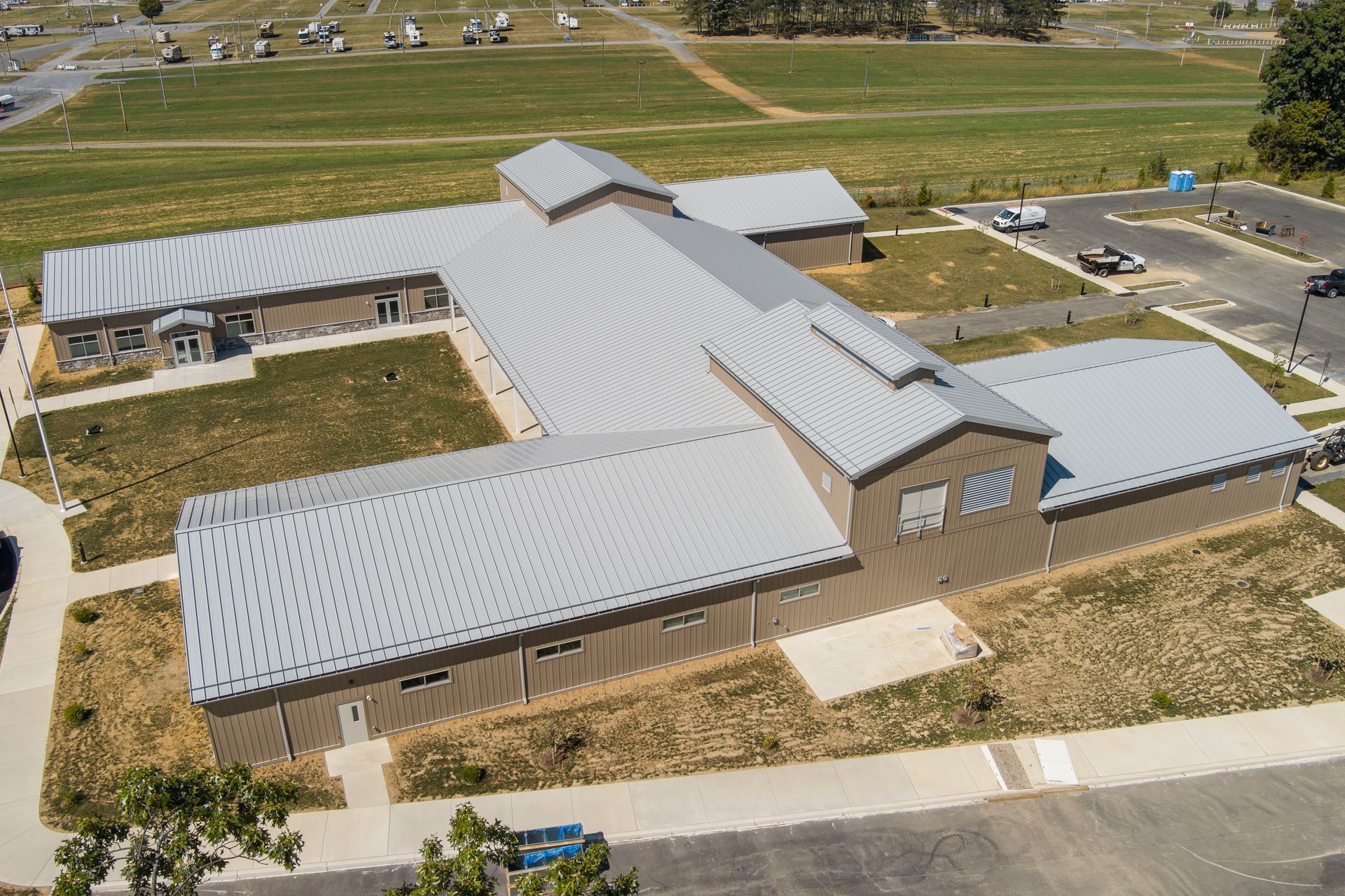 Aerial view of a U-shaped building with a grey metal roof, situated on a grassy landscape with an adjacent parking lot.