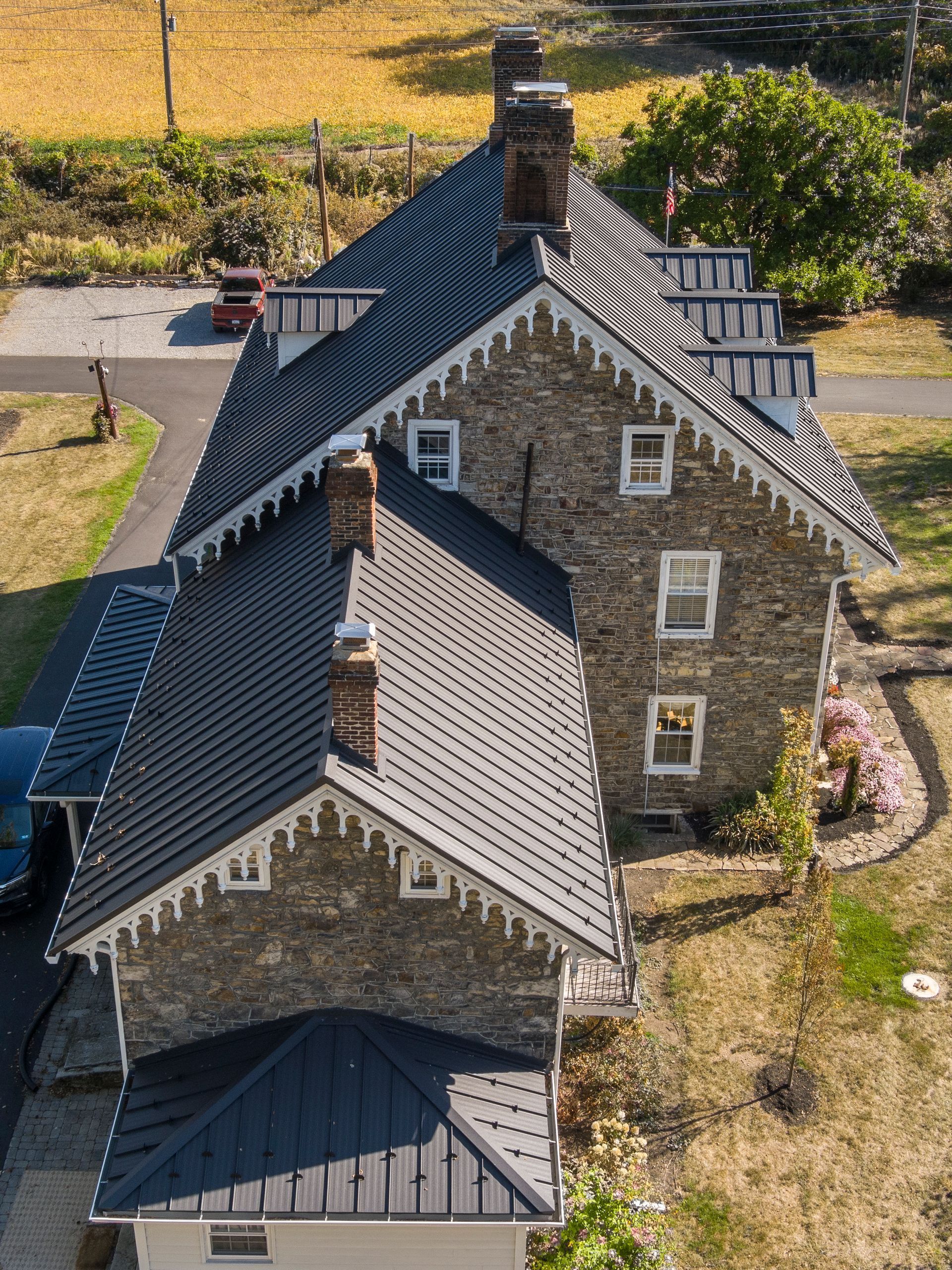 An aerial view of a stone house with a distinctive dark, patterned metal roof and white decorative trim on a sunny day.