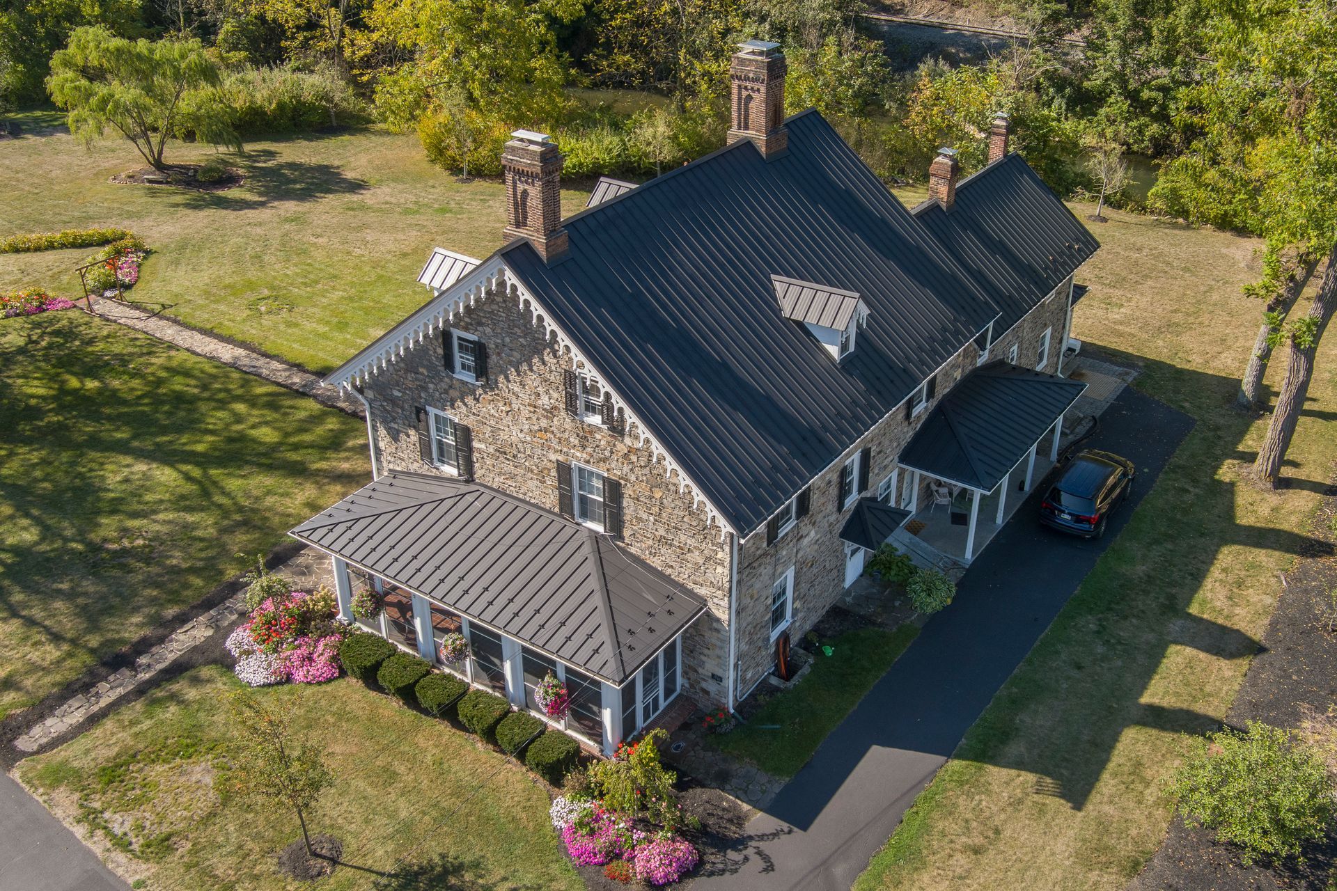 An aerial view of a stone house with a dark metal roof, surrounded by a green lawn, trees, and a paved driveway.