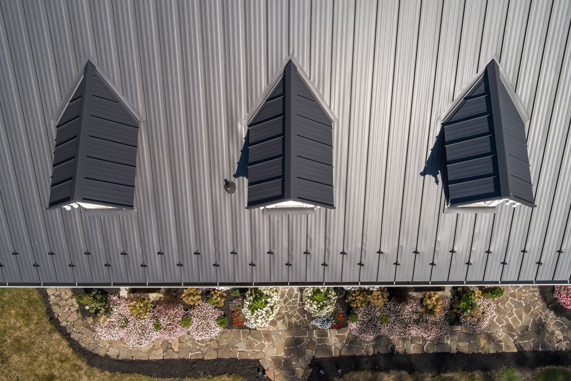 Top-down aerial view of a dark grey metal roof with three gabled dormers above a garden bed with stone paths.