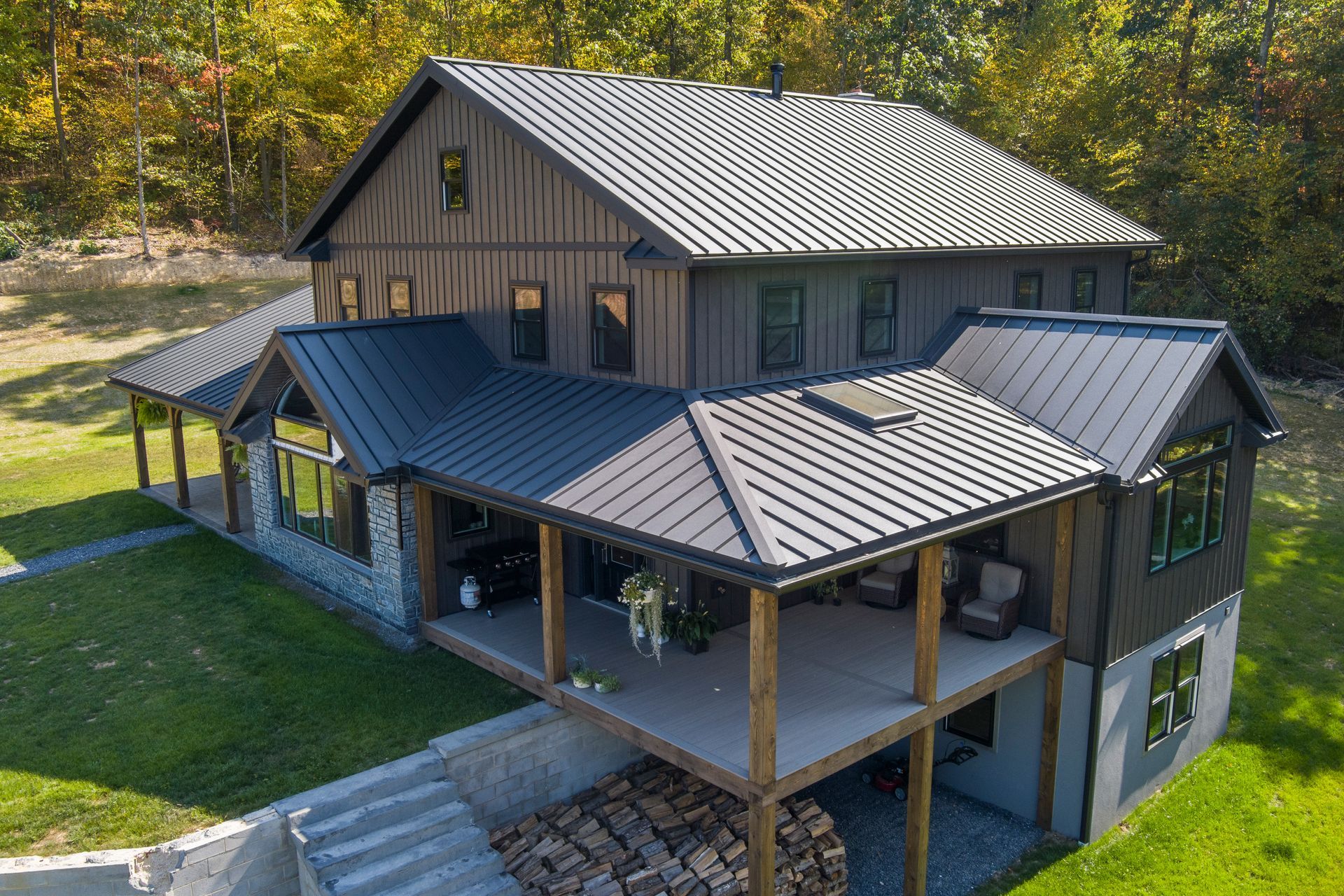 A two-story dark-sided house with a black metal roof, stone foundation, and a wrap-around porch in a wooded yard.