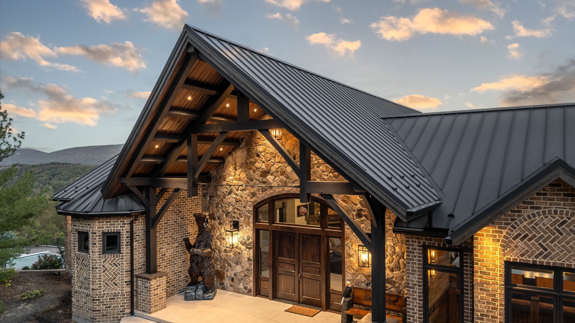 Stone and wood mountain lodge at dusk, with lit windows and a dark metal roof under a cloudy sunset sky