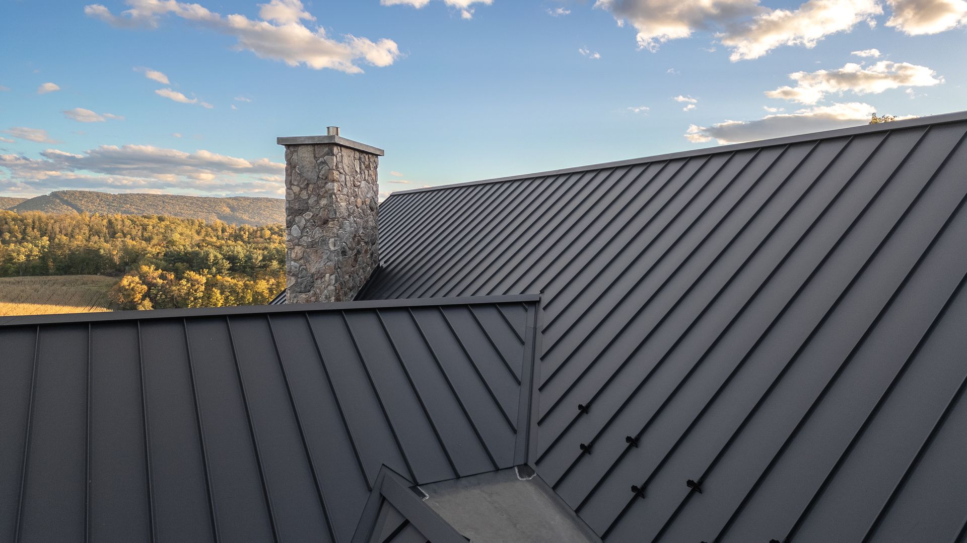 Dark gray metal roof with a stone chimney against a blue sky with clouds, overlooking a forest.