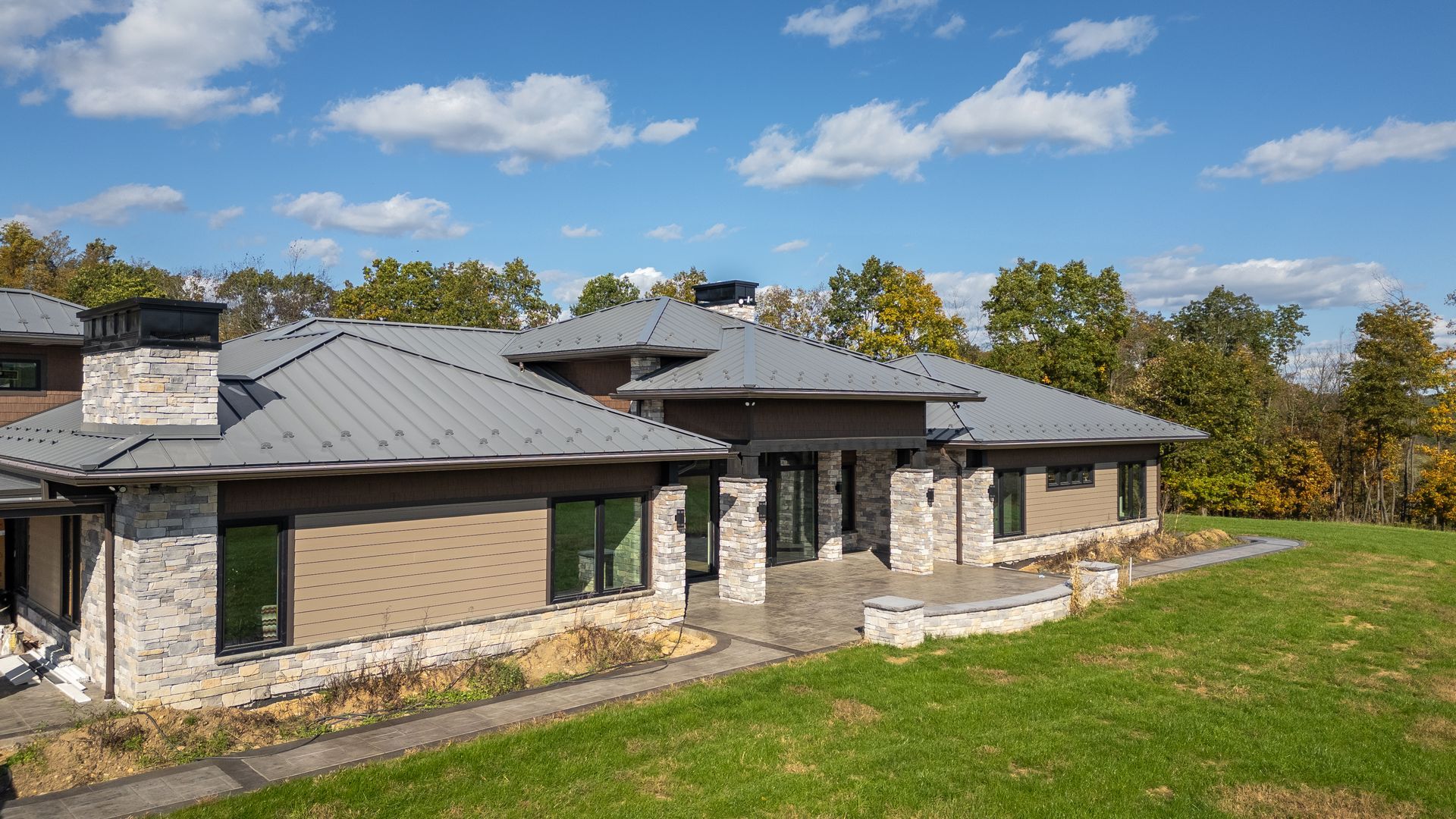 Modern home with gray metal roof, stone and brown siding, on a grassy hill with a blue sky.
