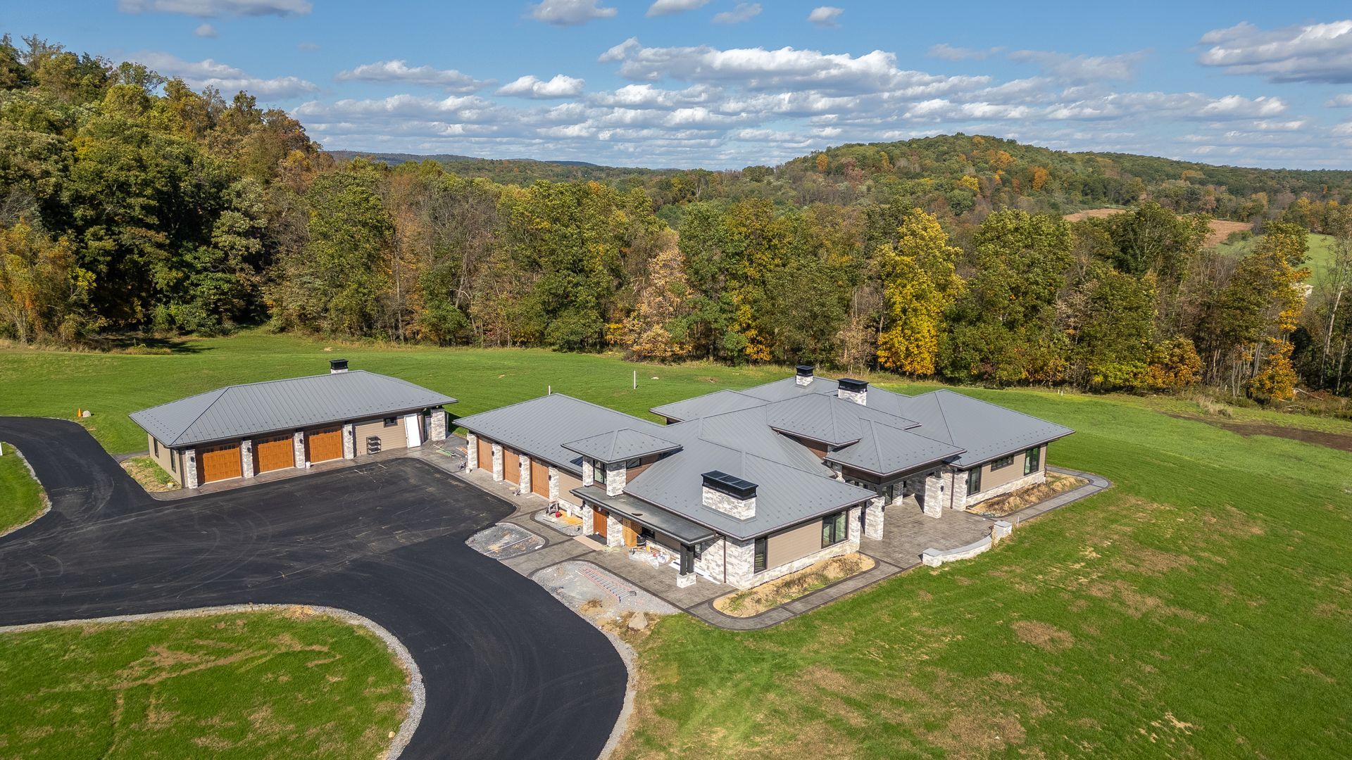 Modern single-story house with attached garage on a green hillside, surrounded by trees. Asphalt driveway.