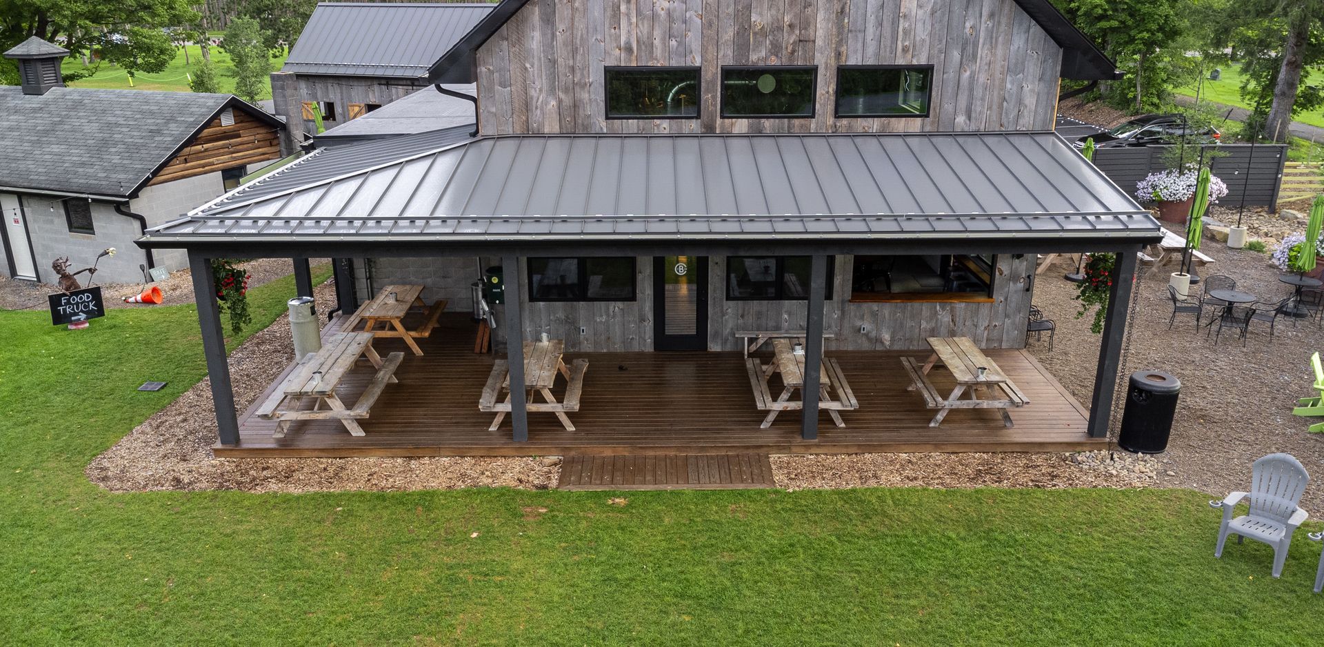 Rustic building with picnic tables on a covered deck, surrounded by green grass and landscaping.